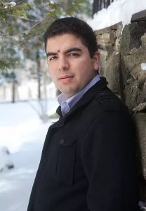 Reinaldo Moya leans against a wall wearing a dark coat and blue collared shirt. Snow and trees are in the background.